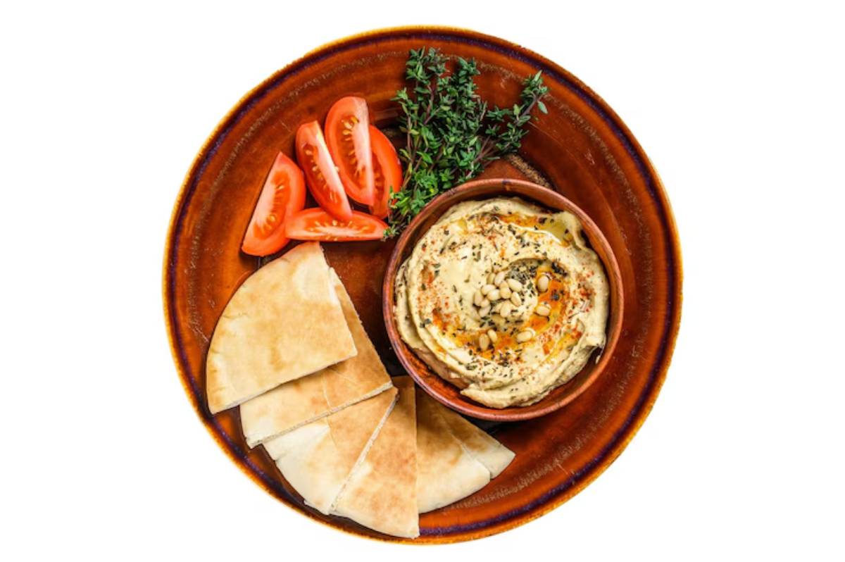 Bowl of hummus topped with pine nuts and herbs, accompanied by sliced tomatoes and pita bread on a rustic plate.