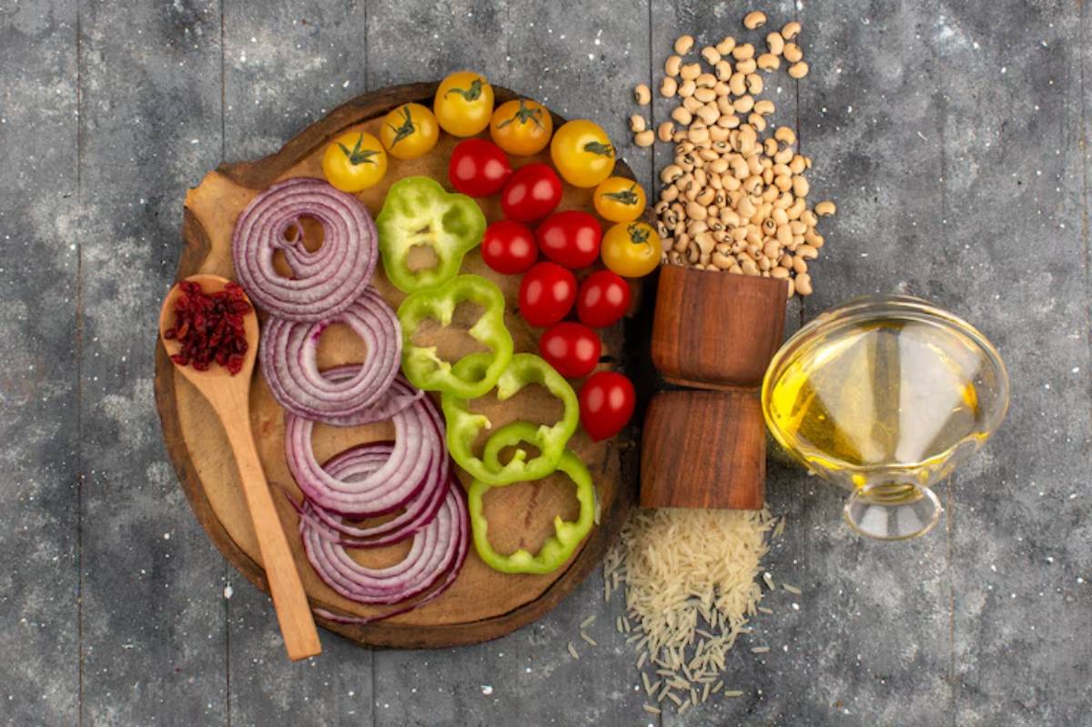 A wooden cutting board with sliced red onions, green bell peppers, cherry tomatoes, yellow tomatoes, rice, black-eyed peas, and olive oil.