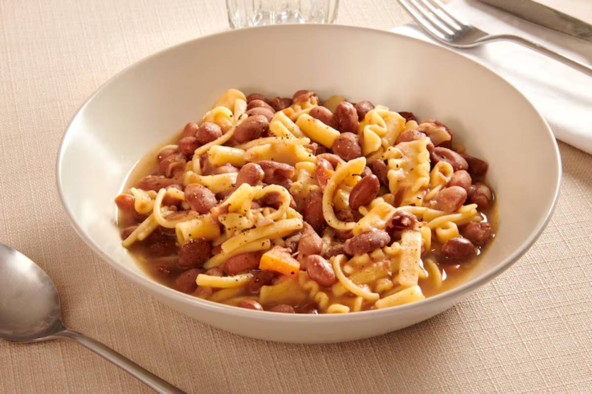 A bowl of pasta with kidney beans, garnished with black pepper, served on a textured beige tablecloth with utensils in the background.