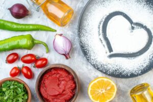 An assortment of fresh ingredients: tomatoes, peppers, onions, herbs, lemon, and oil, with heart-shaped flour design on a black plate.