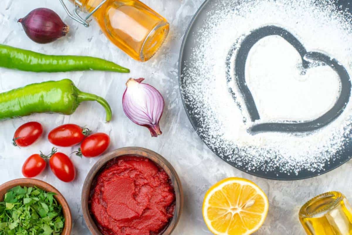 An assortment of fresh ingredients: tomatoes, peppers, onions, herbs, lemon, and oil, with heart-shaped flour design on a black plate.