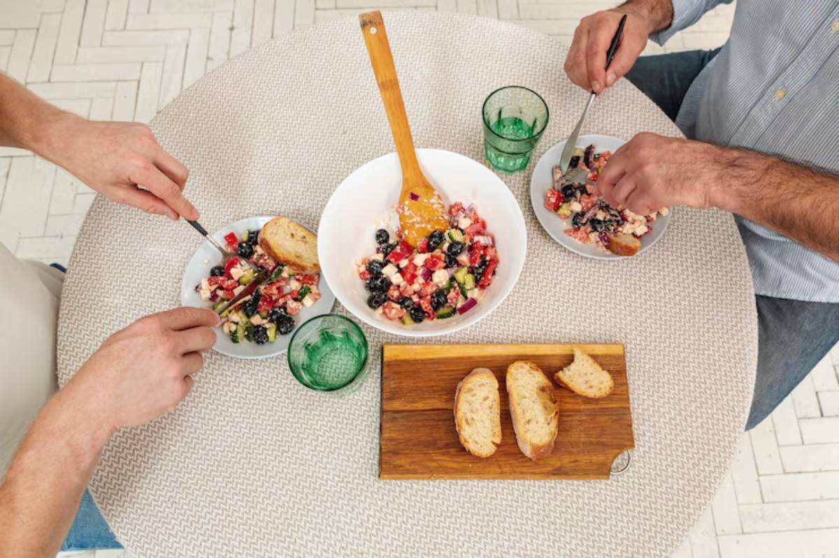 Two people enjoying colorful salads at a round table, with slices of bread and green glasses filled with water.