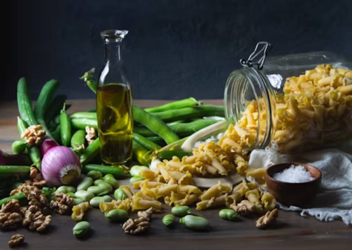 A wooden table showcases spilled pasta, fresh zucchini, fava beans, walnuts, red onion, olive oil, and salt in a rustic setting.