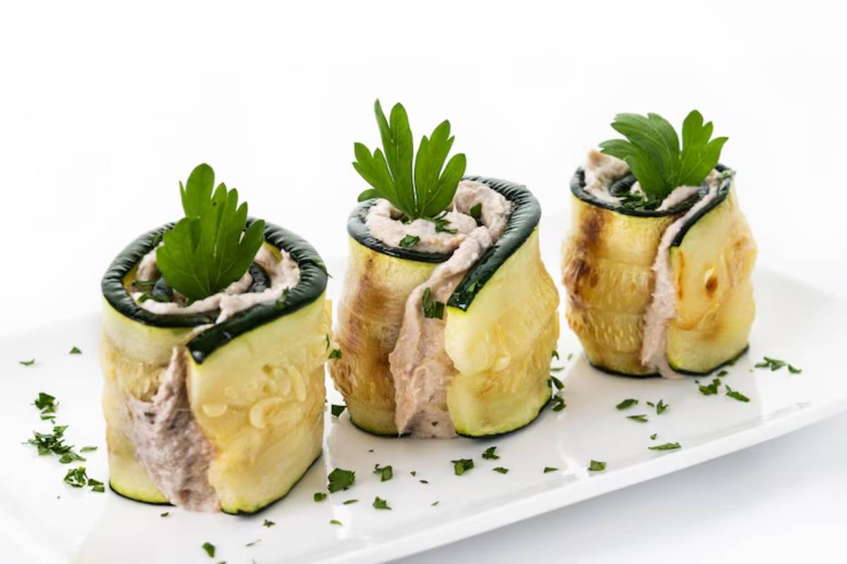 Three elegantly rolled zucchini pieces filled with creamy spread, garnished with fresh parsley, on a white plate.
