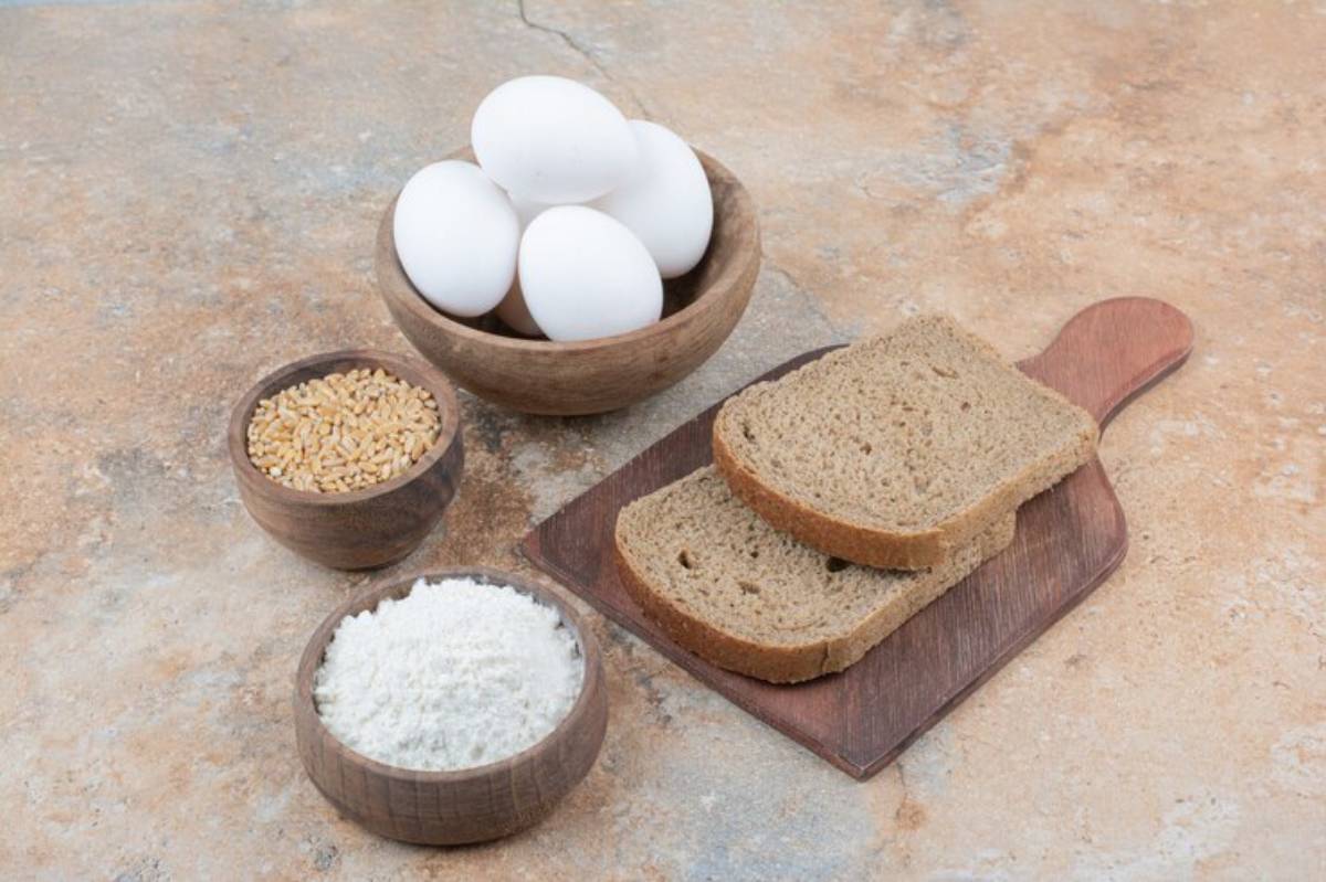 Ingredients for baking: a bowl of eggs, two slices of brown bread, bowls with wheat grains and flour on a textured surface.