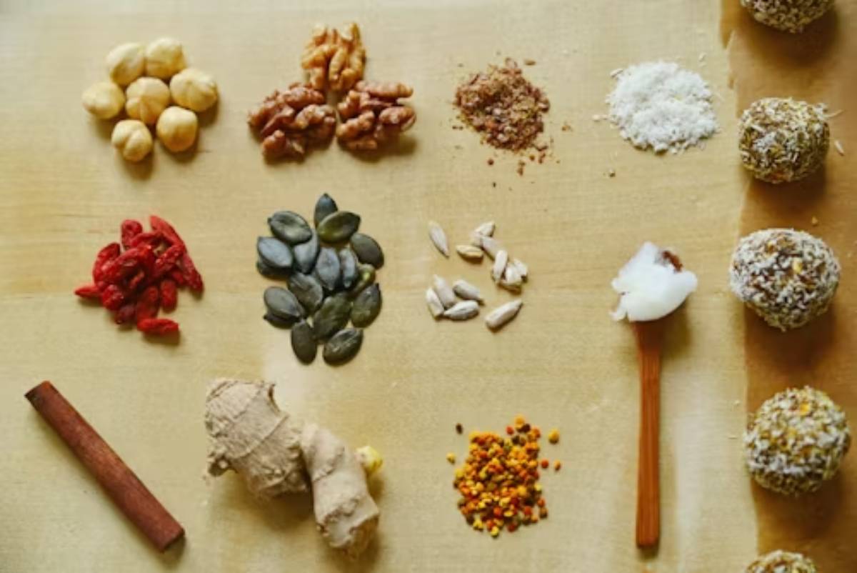 A variety of nuts, seeds, and spices are arranged artistically on a wooden surface, alongside energy balls and a spoonful of coconut.