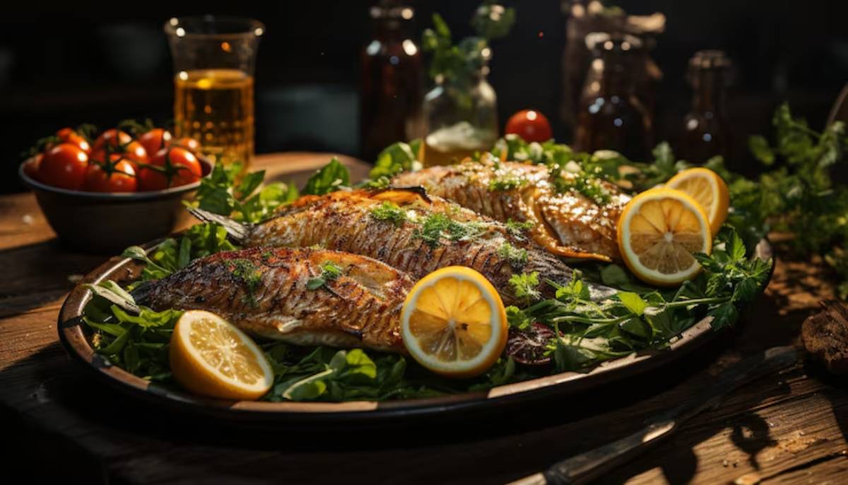 A beautifully arranged plate of grilled fish garnished with lemon slices, fresh greens, and cherry tomatoes, with drinks in the background.