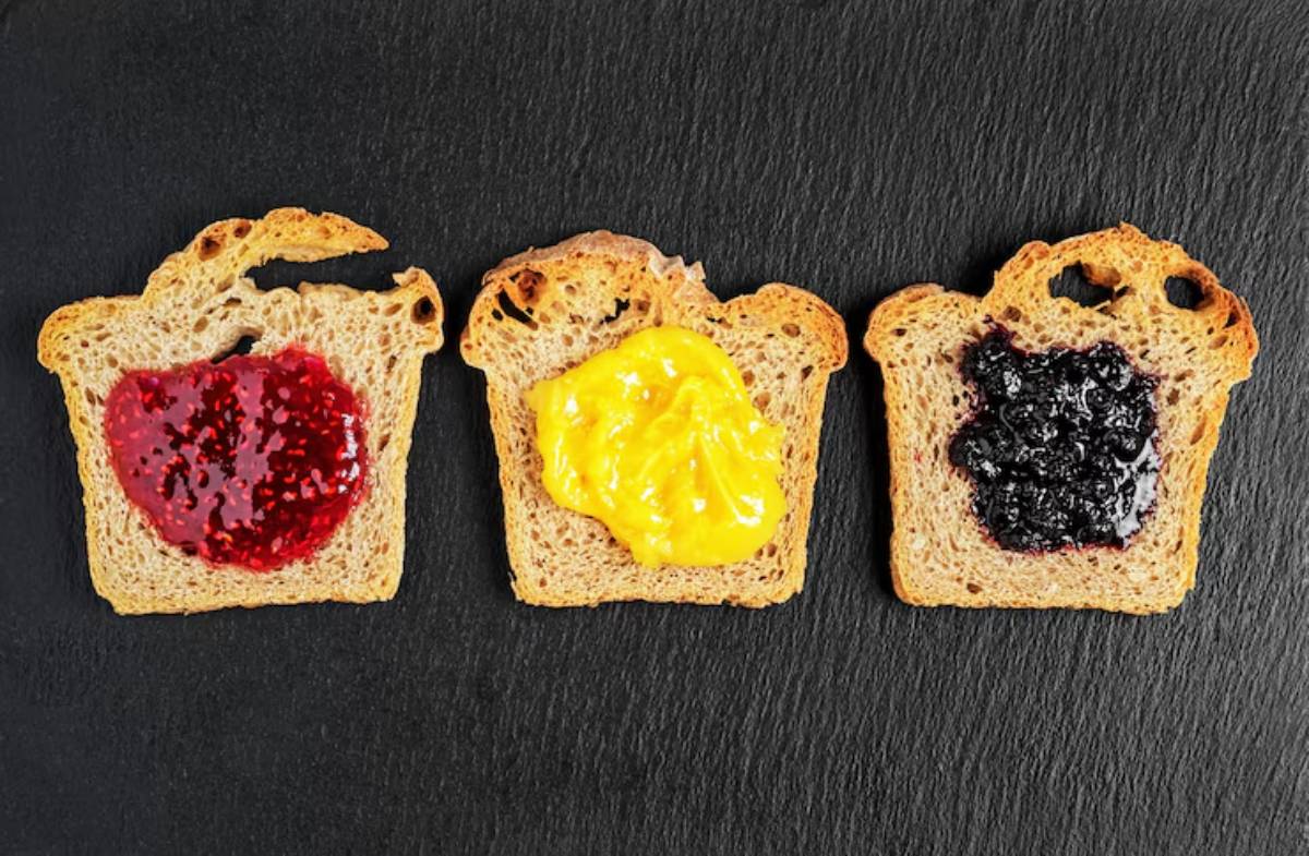 Three slices of toasted bread topped with red jam, yellow mustard, and dark jam, arranged on a black slate surface.