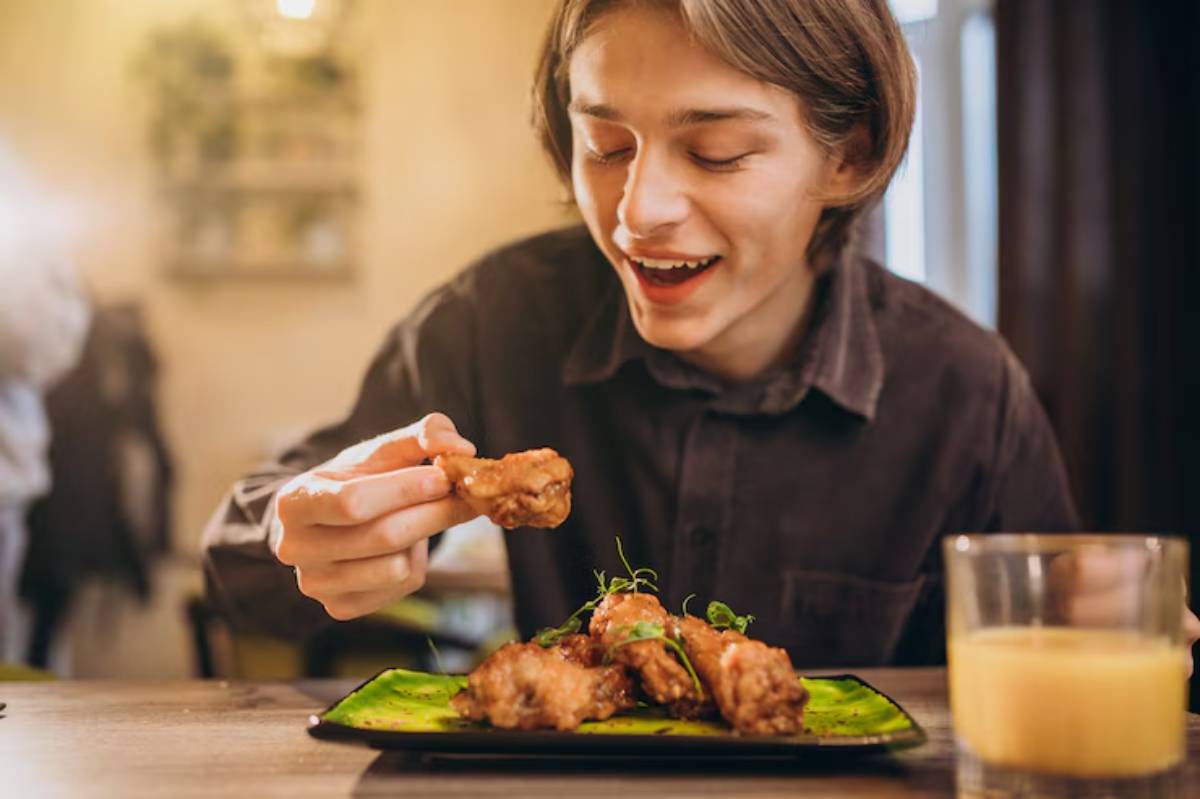 A person holds a piece of fried chicken above a vibrant green plate, accompanied by a glass of juice on a wooden table.