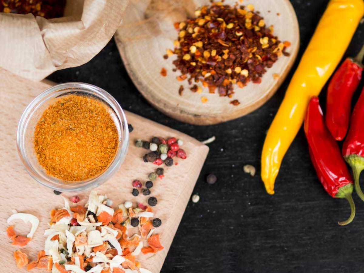 A variety of spices displayed on a wooden board, including a glass jar of orange spice, dried herbs, and colorful chili peppers.