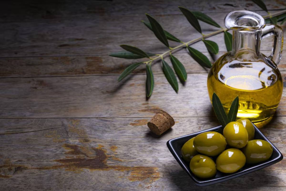 A glass bottle of olive oil beside a bowl of green olives and a cork, with olive leaves on a rustic wooden surface.