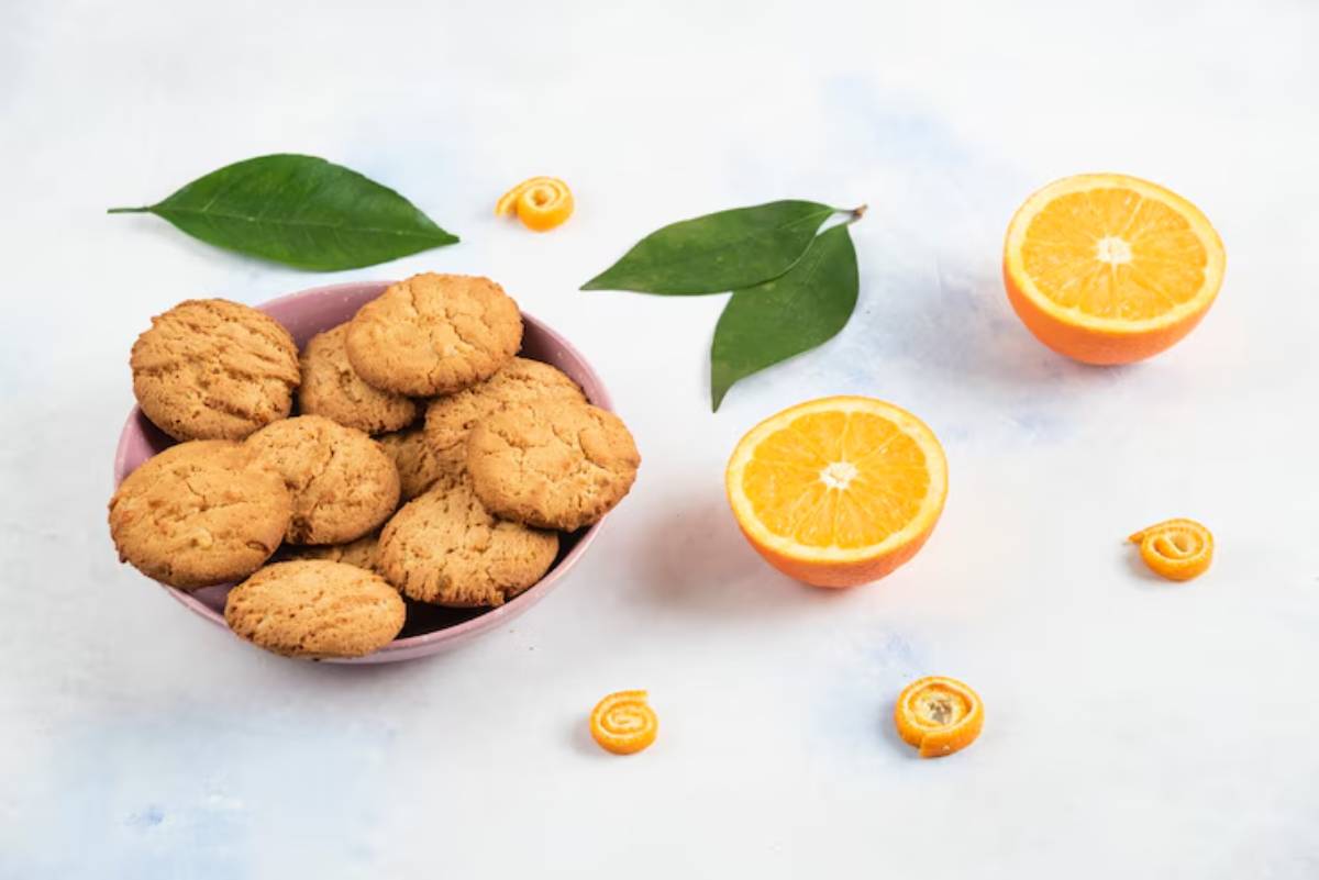A pink bowl filled with golden cookies, surrounded by orange slices, citrus peels, and green leaves on a light background.