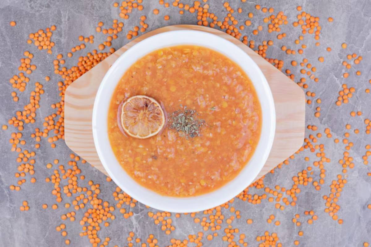 A bowl of orange lentil soup topped with a dried lemon slice and herbs, surrounded by scattered orange lentils on a stone surface.