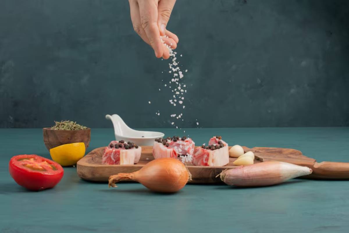 A hand sprinkles salt over sliced meat dishes on a wooden board, surrounded by fresh vegetables and herbs.