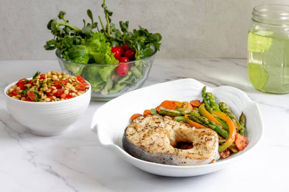 A plate of grilled fish with an assortment of colorful sautéed vegetables, accompanied by a salad and a glass of infused water.