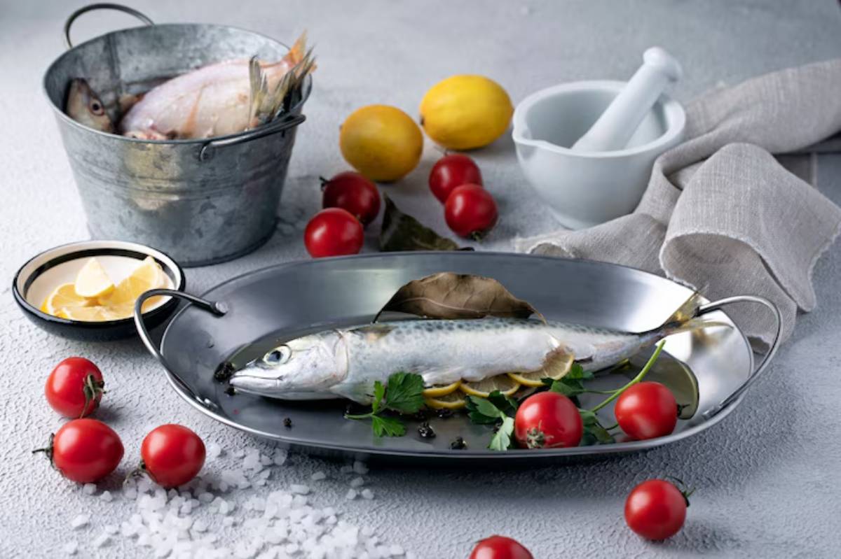 A freshly caught fish rests on a silver platter, surrounded by tomatoes, lemon slices, bay leaves, and a bucket of fish.
