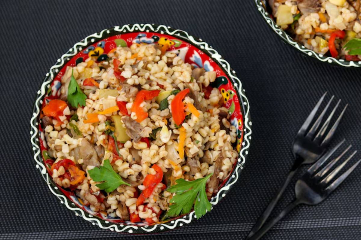 A vibrant bowl of barley mixed with colorful vegetables and meat, garnished with fresh herbs on a textured black background.
