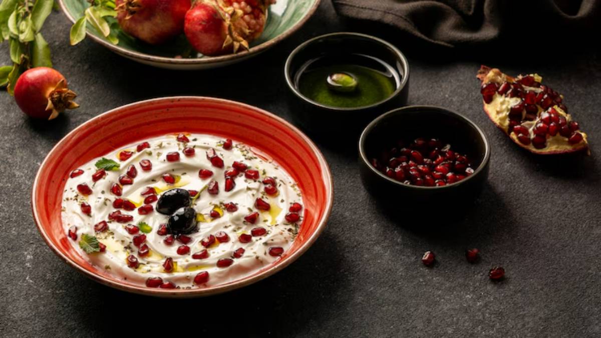 A bowl of yogurt topped with pomegranate seeds and olives, accompanied by pomegranates and olive oil in small dishes on a dark surface.