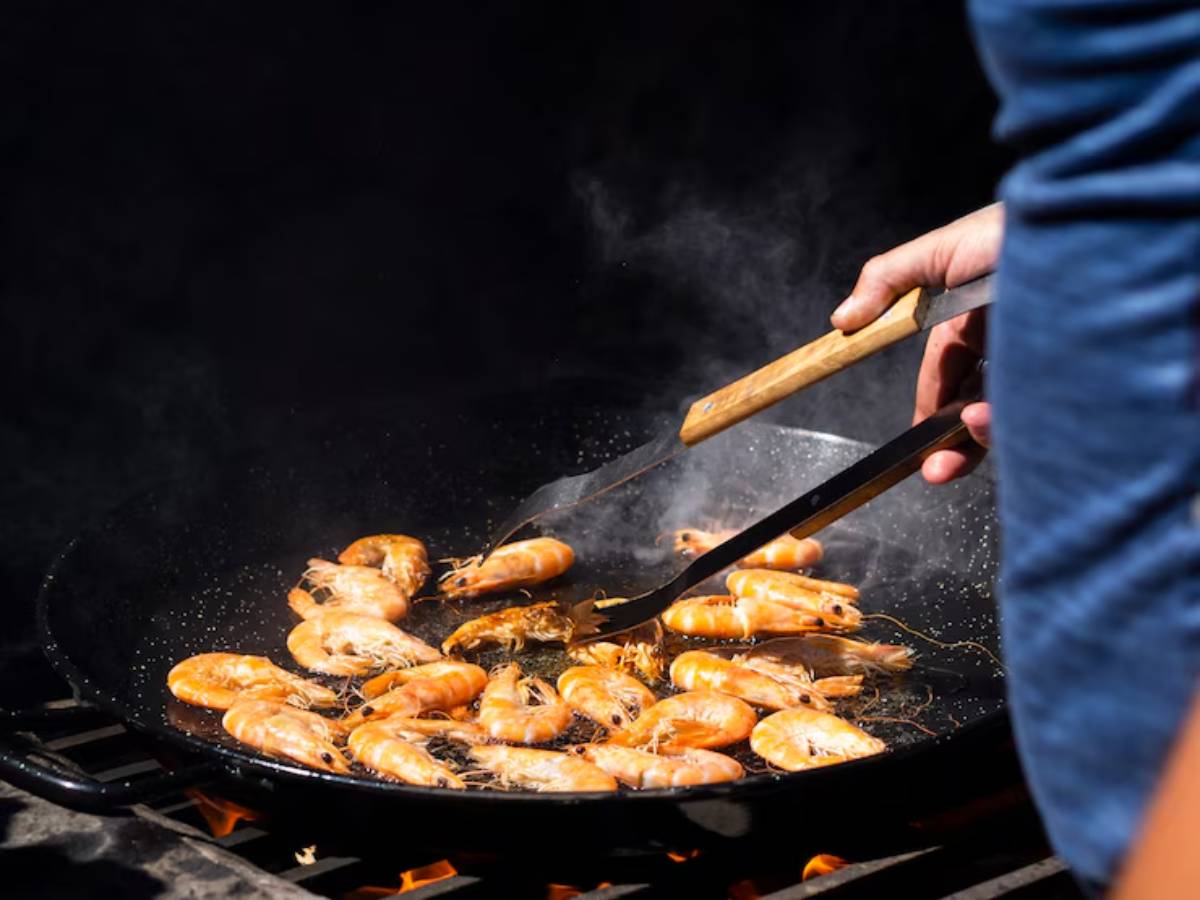 A person cooks shrimp in a large skillet over an open flame, using tongs to flip the seafood as steam rises around them.
