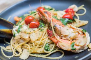 A close-up of spaghetti topped with large shrimp, cherry tomatoes, sliced garlic, and green herbs on a black plate.