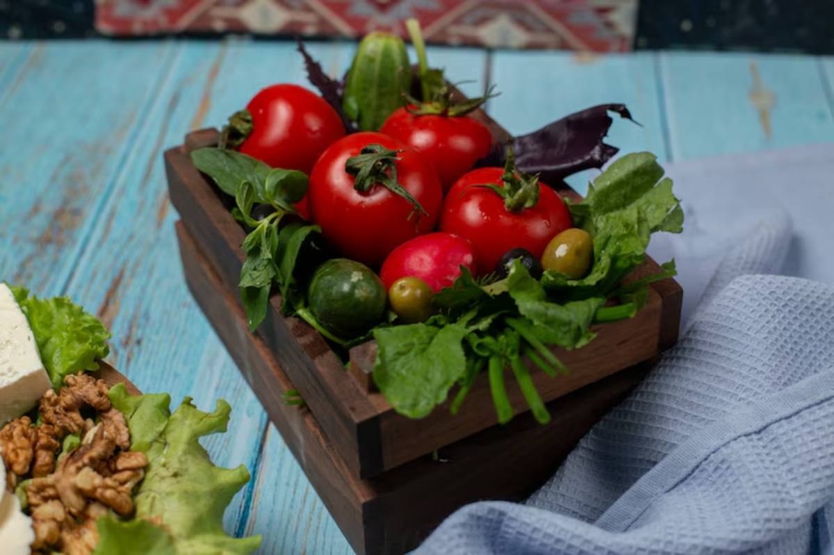 A wooden crate holds fresh red tomatoes, greens, and olives, set against a light blue wooden background and a textured cloth.