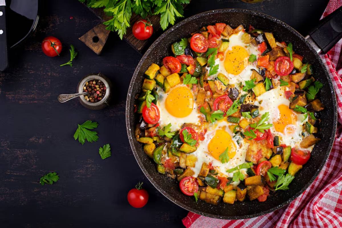 A vibrant skillet dish with sunny-side-up eggs, tomatoes, zucchini, and herbs, surrounded by a red and white checkered cloth.
