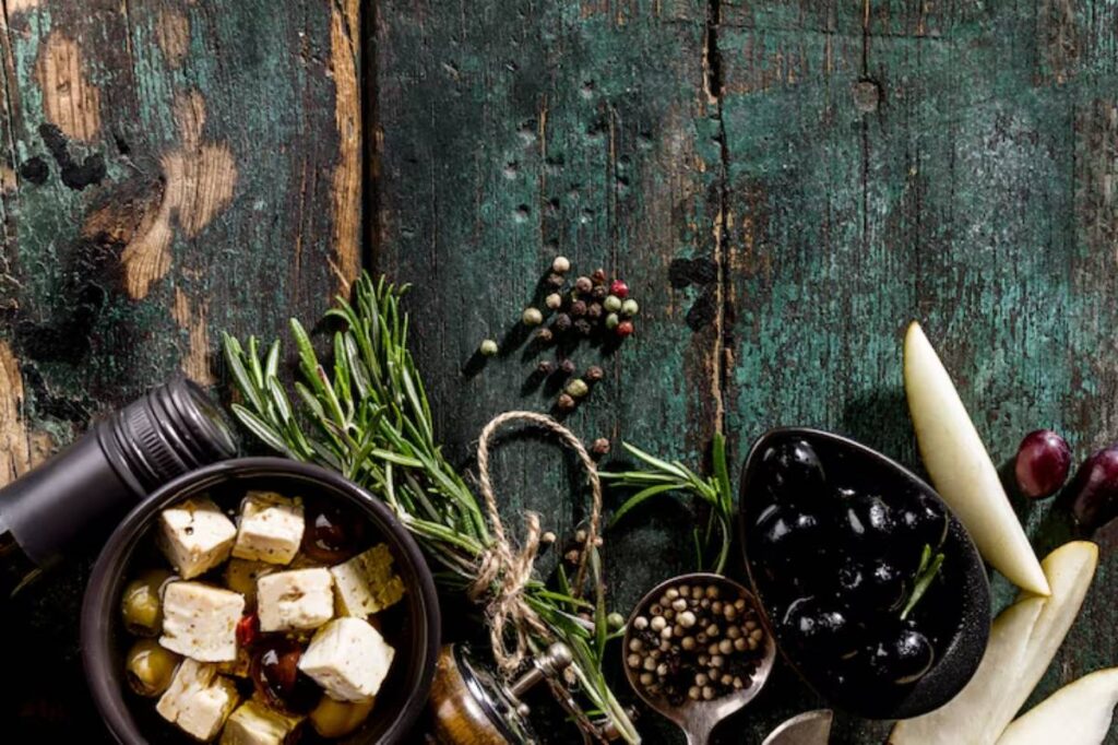 An array of olives, herbs, spices, and cheese cubes on a rustic wooden table, showcasing vibrant colors and textures.
