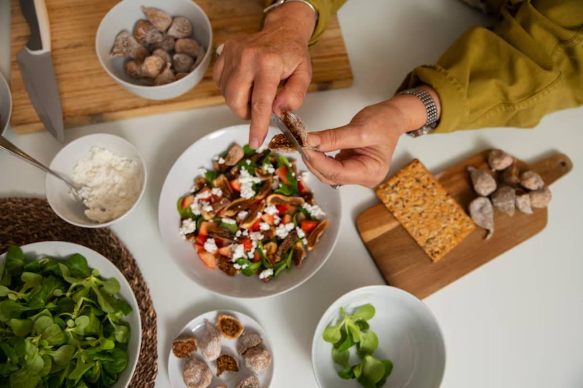 A person prepares food, pulling apart a dried fig, surrounded by fresh salad, cheese, and snacks on a white table.