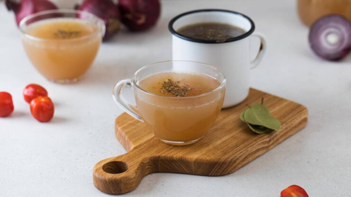 Two cups of broth on a wooden board, with cherry tomatoes and bay leaves nearby, set against a light background with onions.
