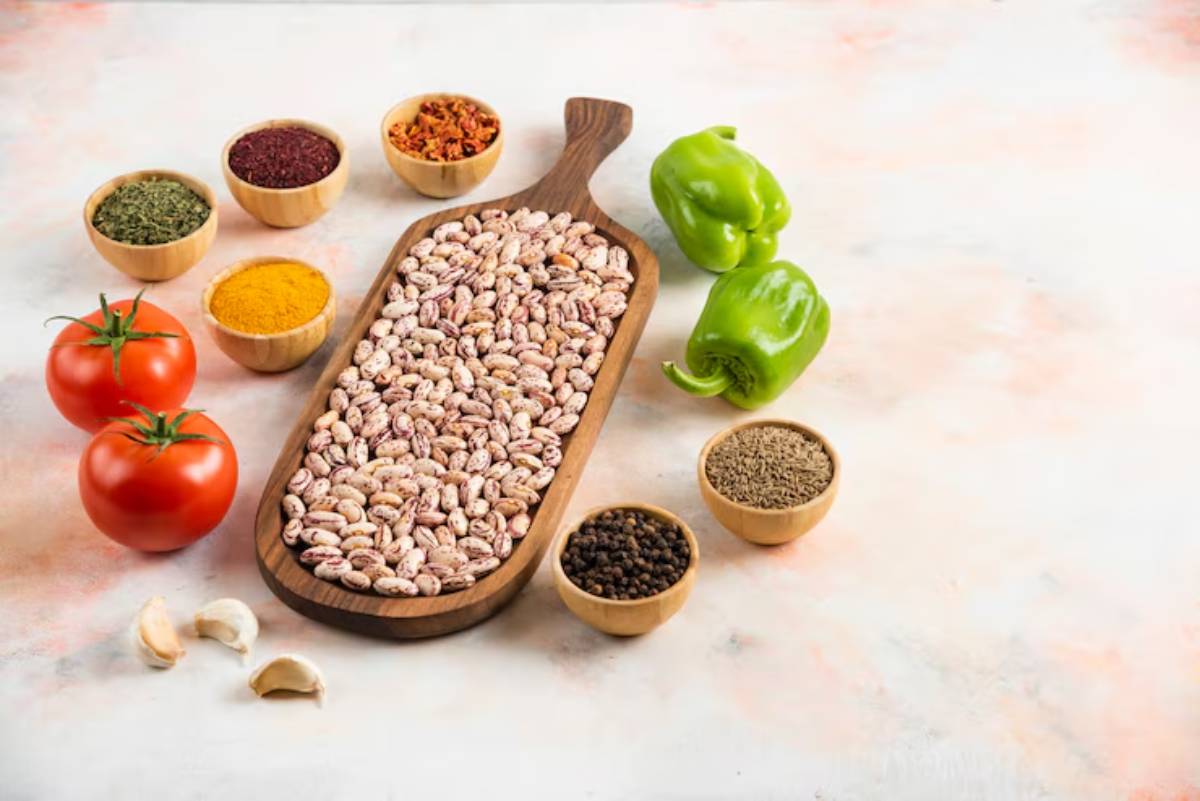 A wooden platter holds various dried beans, surrounded by bowls of spices and vibrant green bell peppers on a soft, pastel surface.
