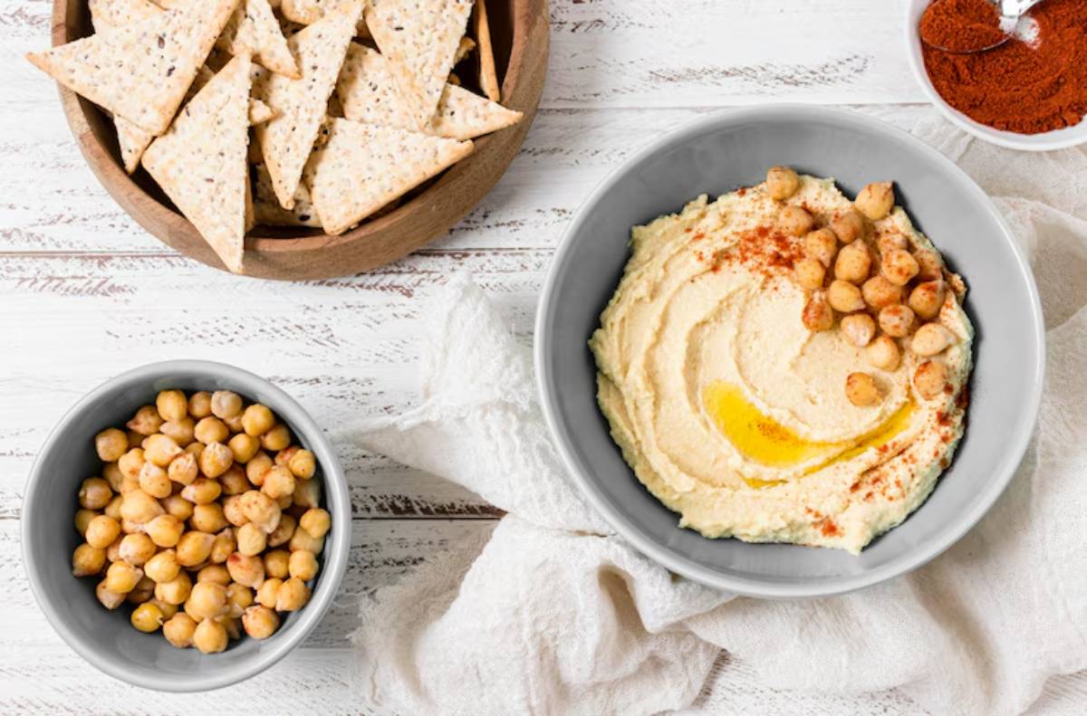 A bowl of creamy hummus topped with chickpeas and olive oil, accompanied by crispy pita chips and a small bowl of spice.