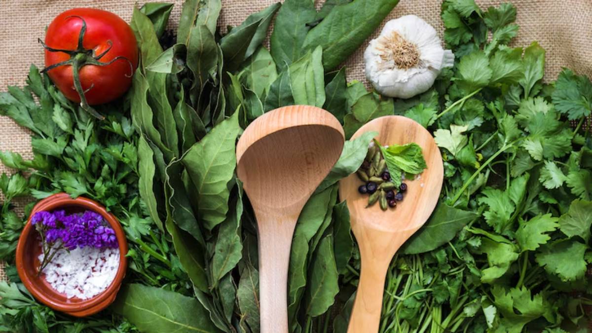 A vibrant arrangement of fresh herbs, a tomato, garlic, wooden spoons, and seasoning on a textured fabric background.