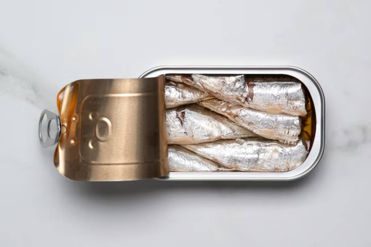Open metal can of sardines, showcasing several silver-skinned fish resting in golden oil against a white marble background.