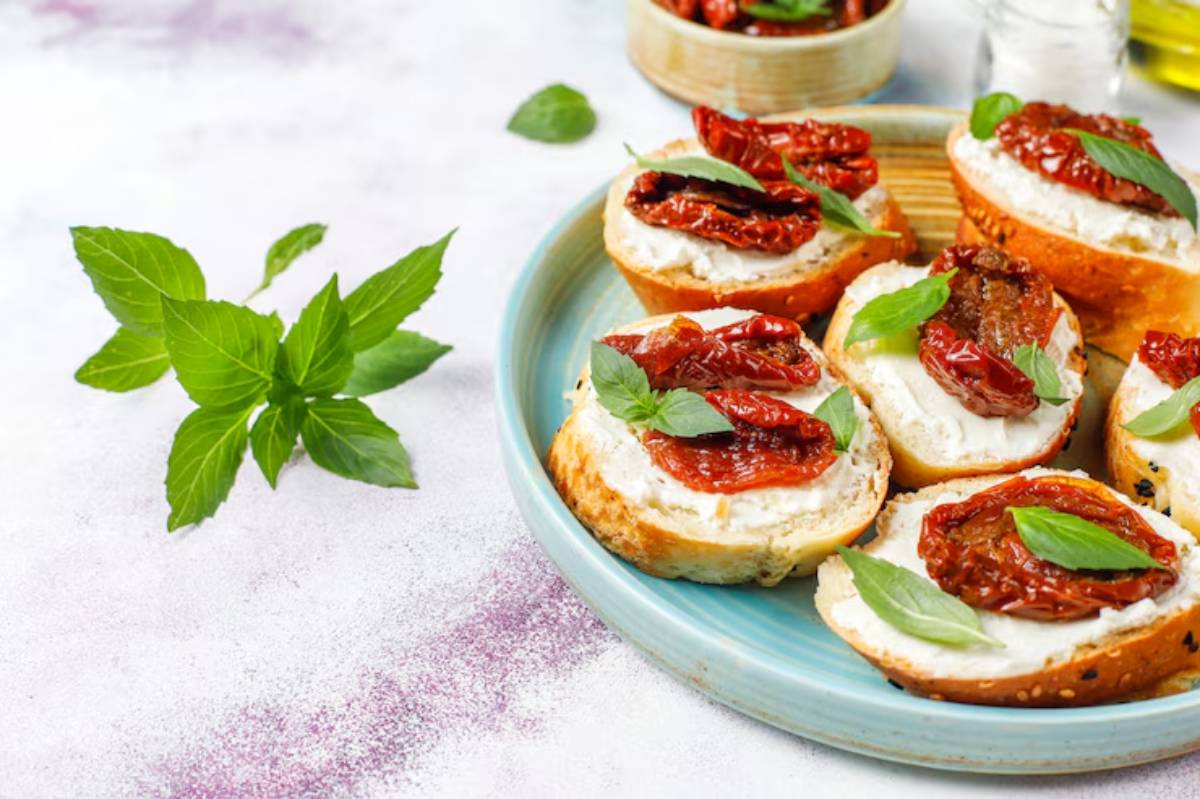 A plate of toasted bread topped with creamy cheese, sun-dried tomatoes, and fresh basil leaves, with a sprig of basil nearby.