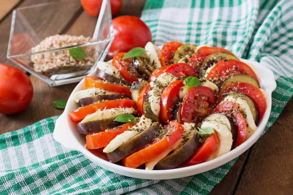 A colorful ratatouille dish with layers of sliced tomatoes, eggplant, and cheese, garnished with herbs on a checkered cloth.