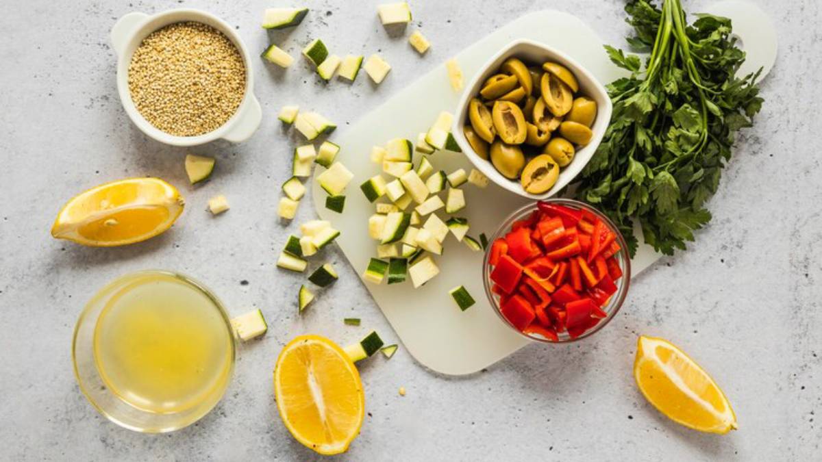 A flat lay of fresh ingredients: chopped zucchini, red pepper, olives, quinoa, lemon wedges, and cilantro on a light background.