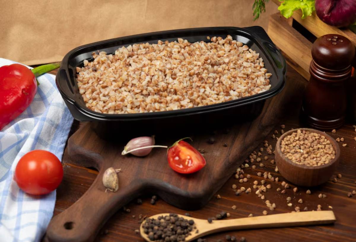 A black container filled with cooked buckwheat, surrounded by fresh vegetables and spices on a wooden surface.