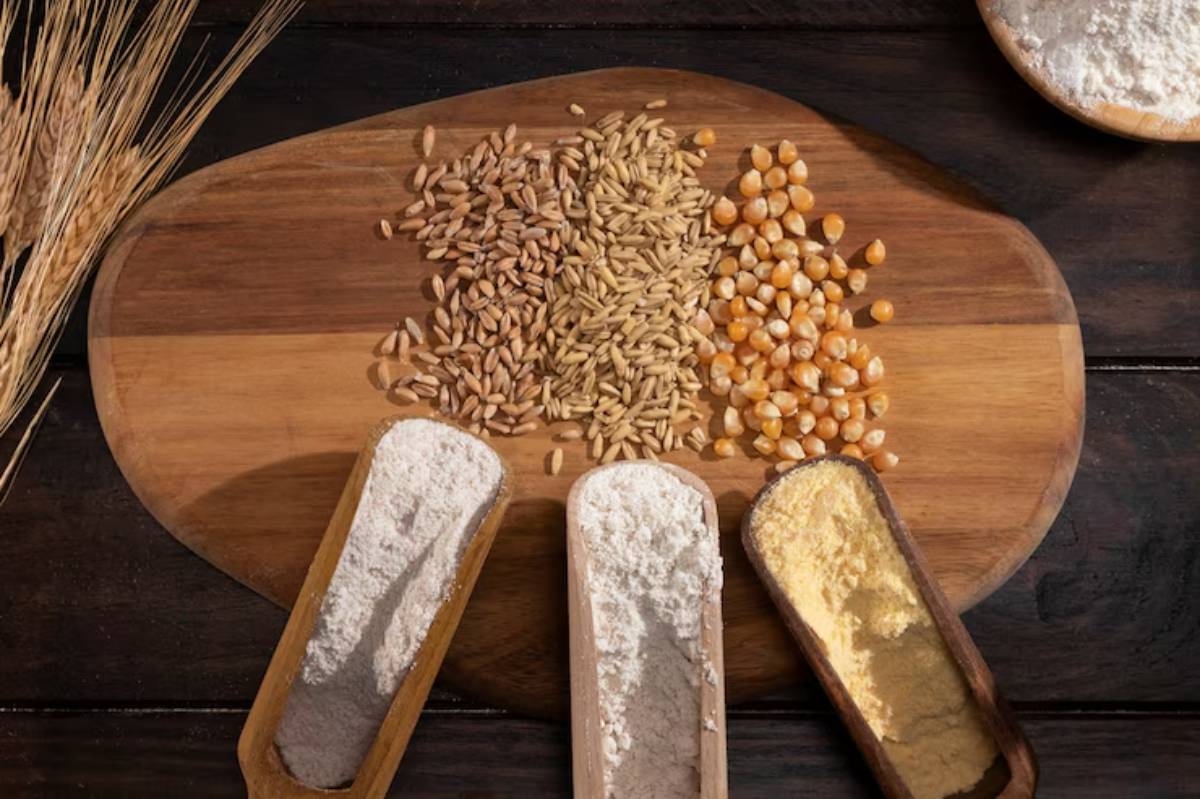A wooden platter displays various grains and flours: wheat, rye, corn kernels, and three scoops of different flours.