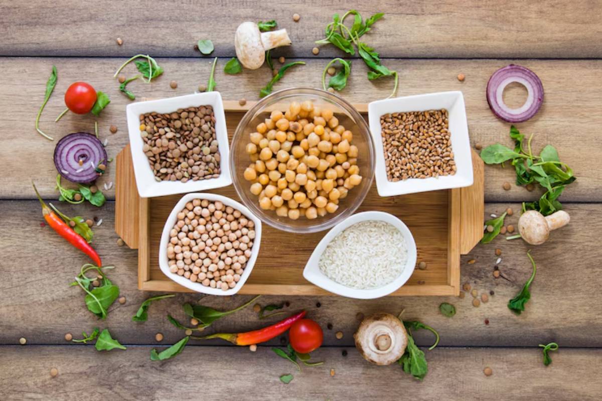 A wooden tray displays bowls of chickpeas, lentils, mung beans, rice, and wheat surrounded by fresh vegetables and herbs on a rustic table.