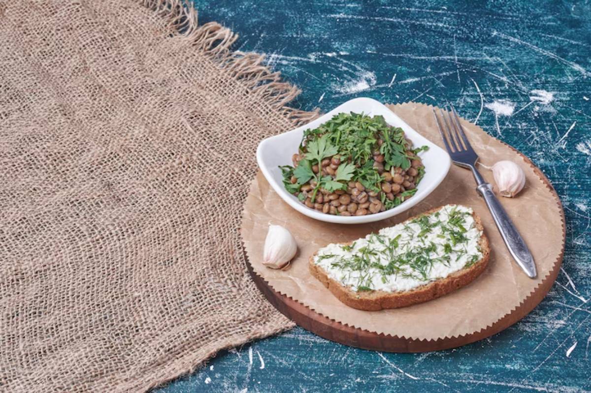A rustic wooden plate holds a slice of bread topped with cream and herbs, alongside lentils with greens and a garlic clove.