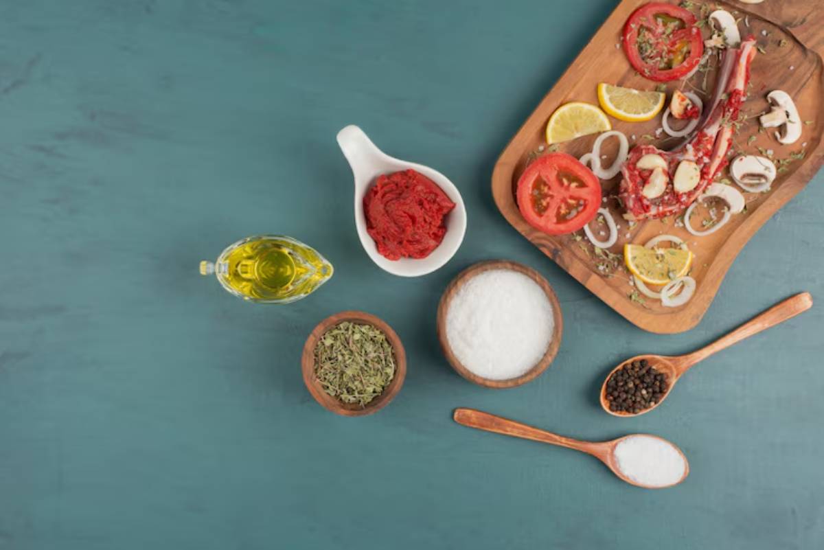 A wooden platter with fresh tomatoes, lemons, onions, and mushrooms, alongside olive oil, spices, and tomato paste on a teal background.