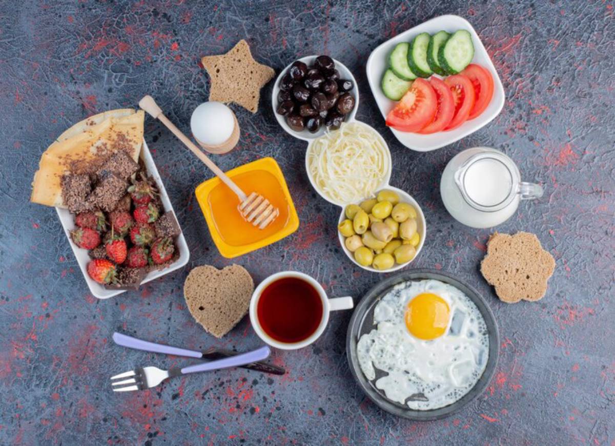 A colorful breakfast spread featuring fresh fruits, vegetables, olives, cheese, honey, eggs, tea, and bread on a textured background.