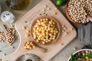A wooden board displays cooked chickpeas in a bowl, surrounded by raw chickpeas, salt, pepper, cucumber, and olive oil.