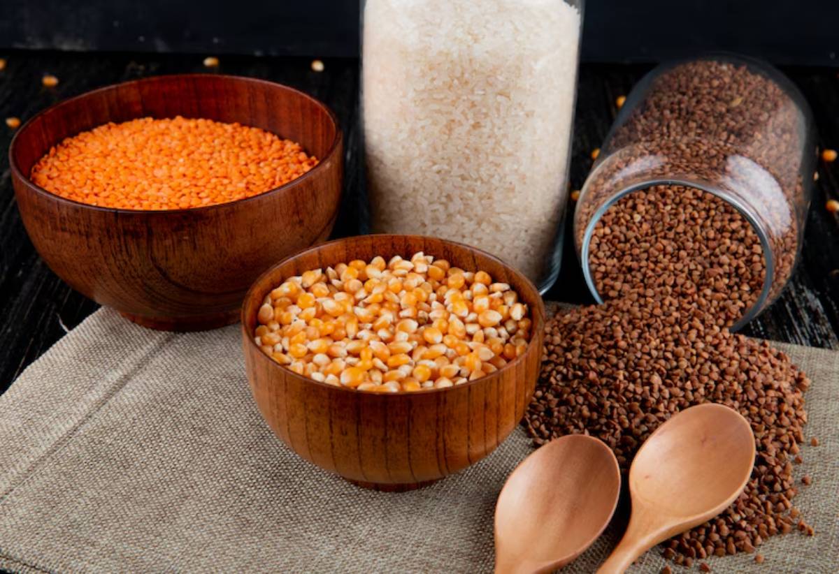 A wooden table displays three bowls of different grains: orange lentils, yellow peas, and brown grains, with a jar of rice in the background.