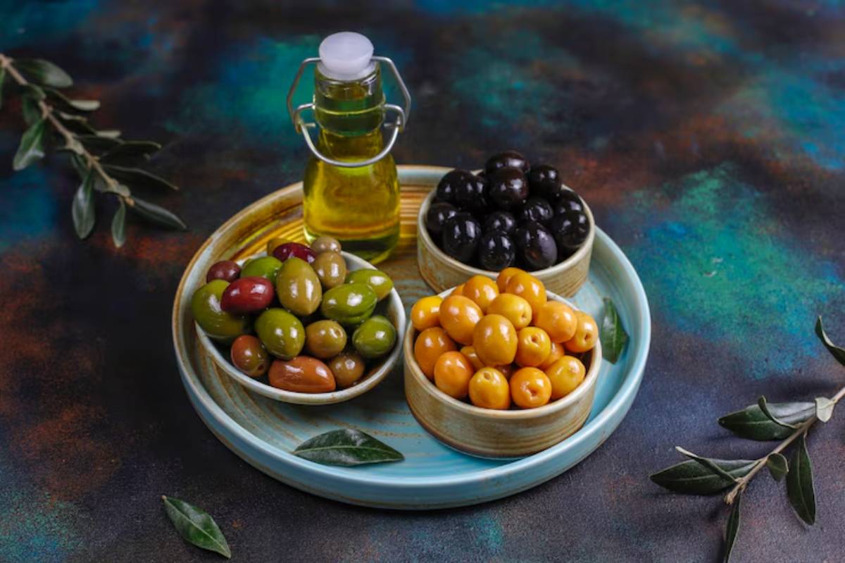 A colorful display of various olives in bowls, with a bottle of olive oil, set against a vibrant background.