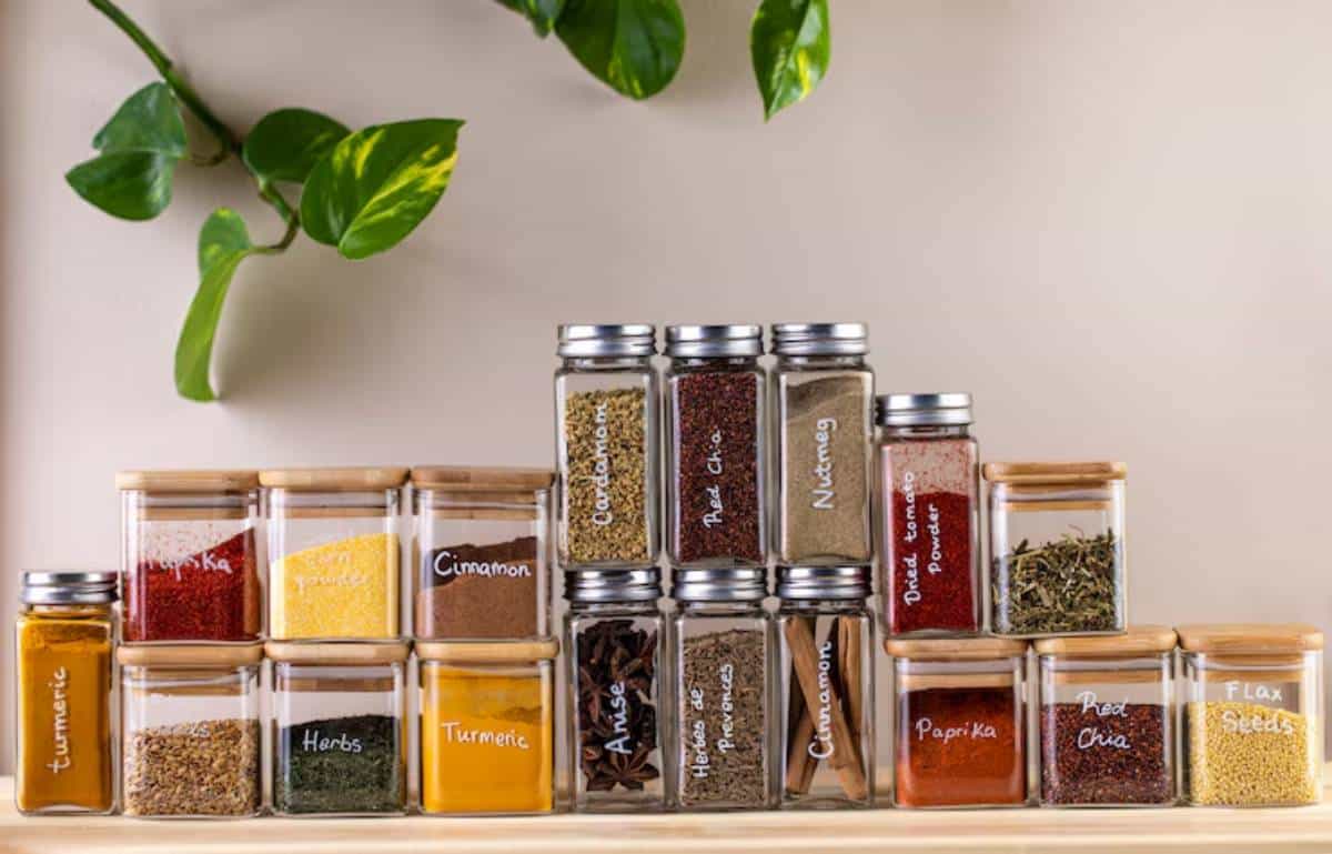 A neat display of labeled spice jars on a wooden shelf, featuring vibrant spices like turmeric, paprika, and cinnamon, with leafy greenery above.