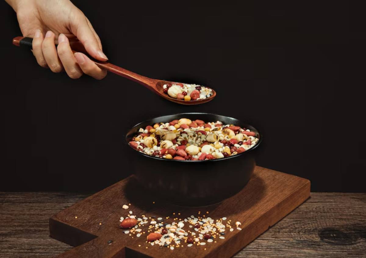A hand scoops a variety of colorful grains and nuts from a wooden bowl with a spoon onto a dark backdrop.