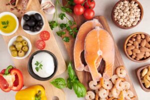 A colorful array of fresh ingredients including salmon, shrimp, tomatoes, olives, herbs, and nuts on a wooden board.