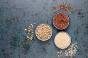 Three bowls filled with various seeds: pine nuts, flax seeds, and sesame seeds, displayed on a textured green surface.