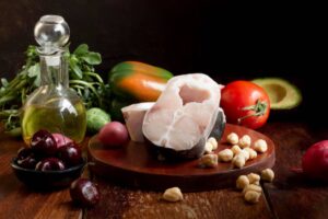 A wooden board displays fresh fish, olive oil, assorted vegetables, cherries, and chickpeas, creating a vibrant, healthy scene.